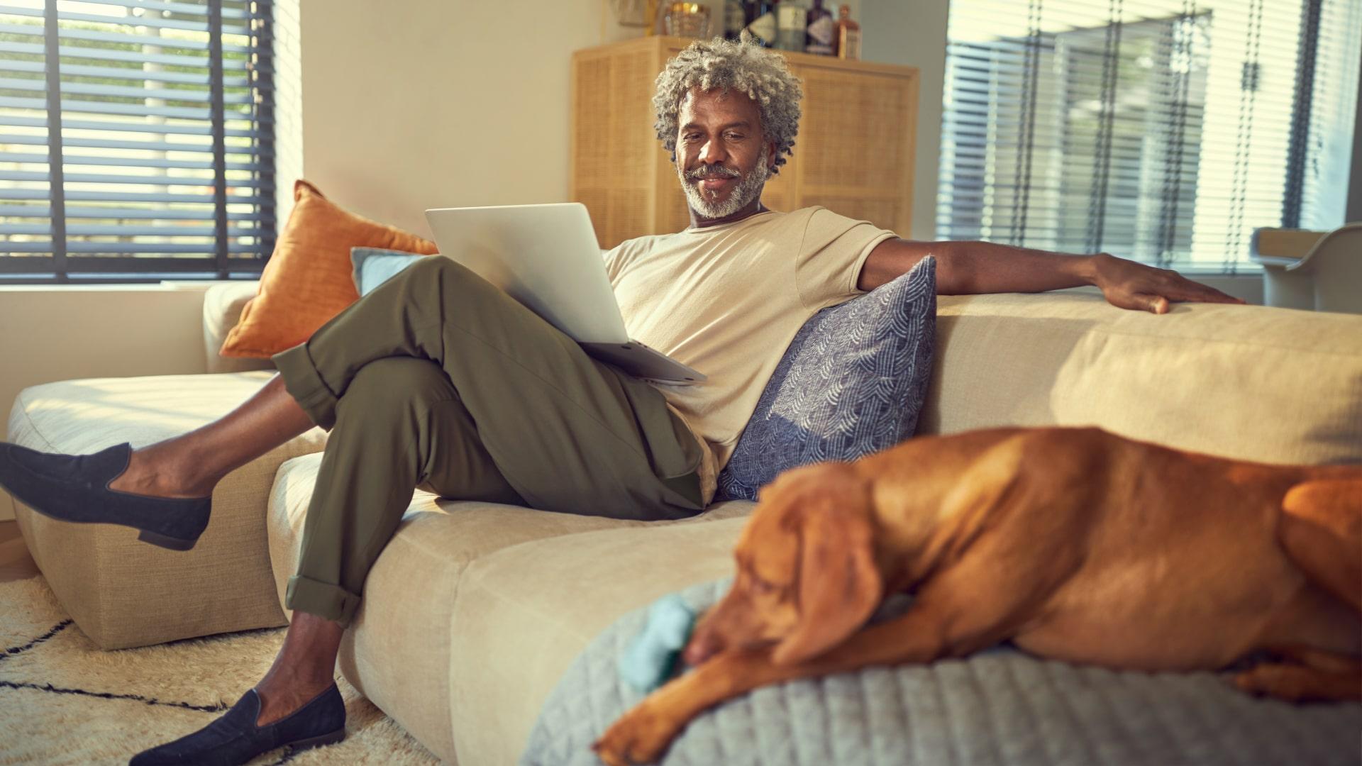 Homem sentado no sofá trabalhando no seu laptop com o seu cão deitado ao lado