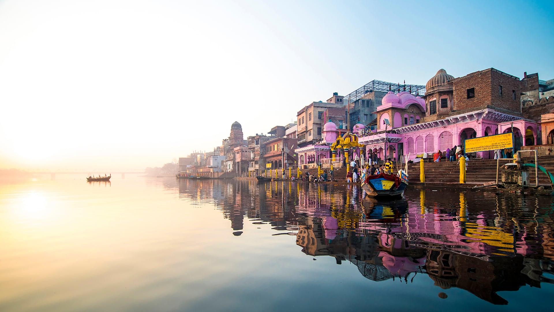 Sunrise over river with reflections of colorful buildings and a boat in India.