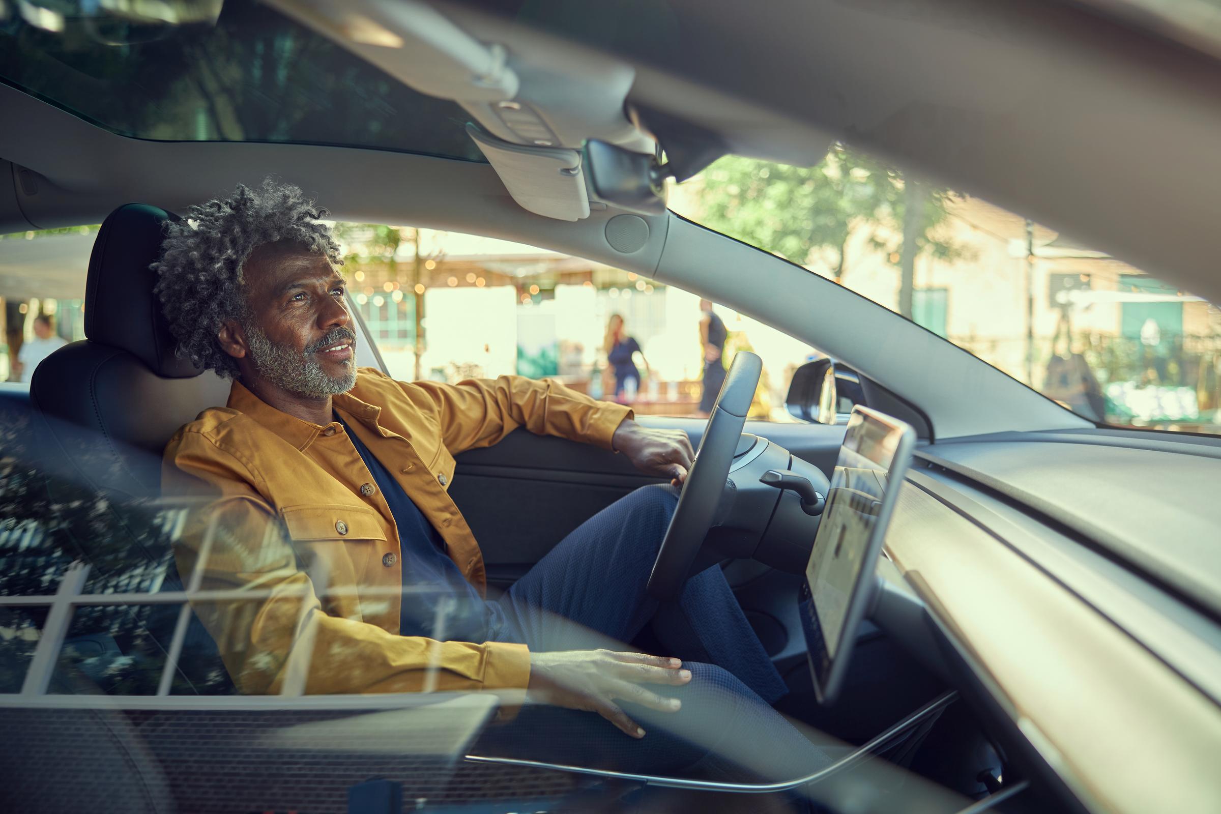 Homem maduro com cabelos grisalhos sorrindo enquanto senta no banco do motorista de um carro moderno.