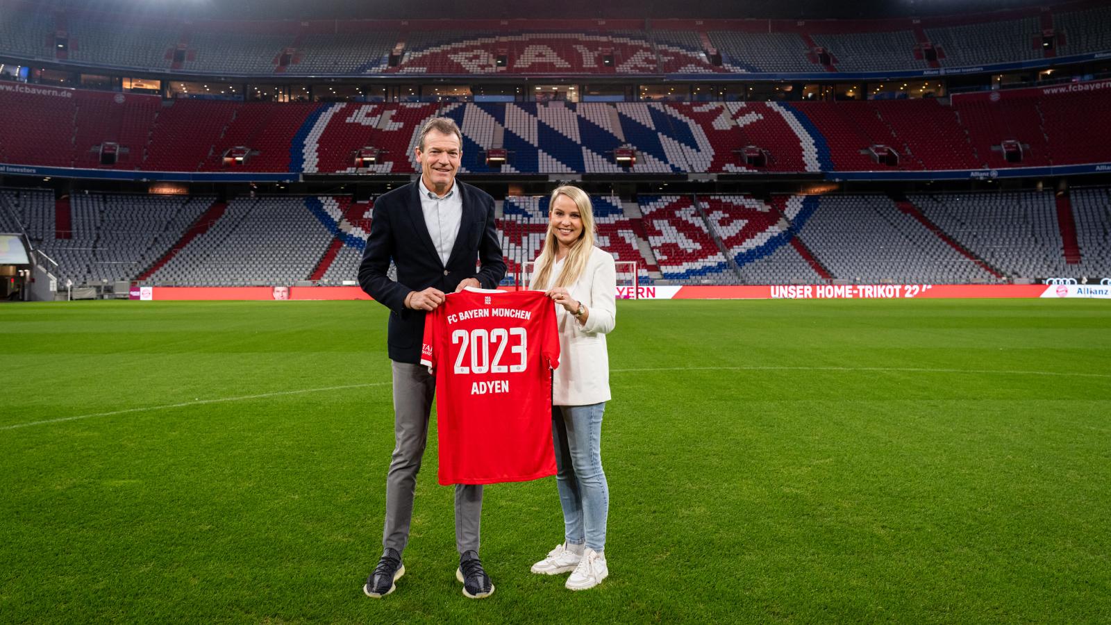 Two individuals holding a red FC Bayern Munchen jersey with '2023 Adyen' in a stadium.