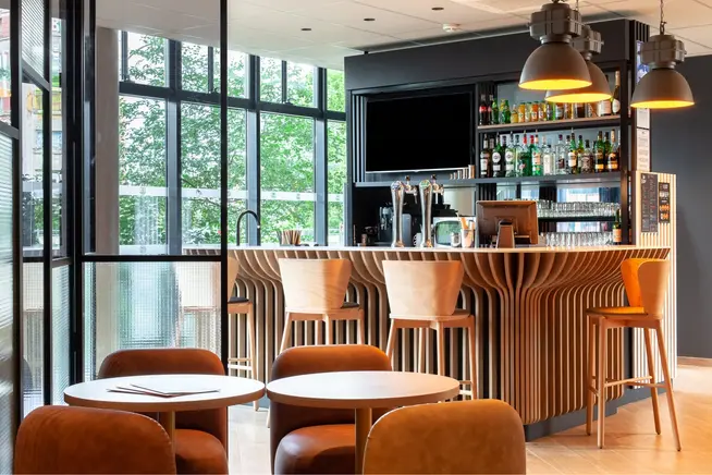 Interior de cafeteria moderna com balcão de madeira, bancos e mesas rodeadas por grandes janelas.