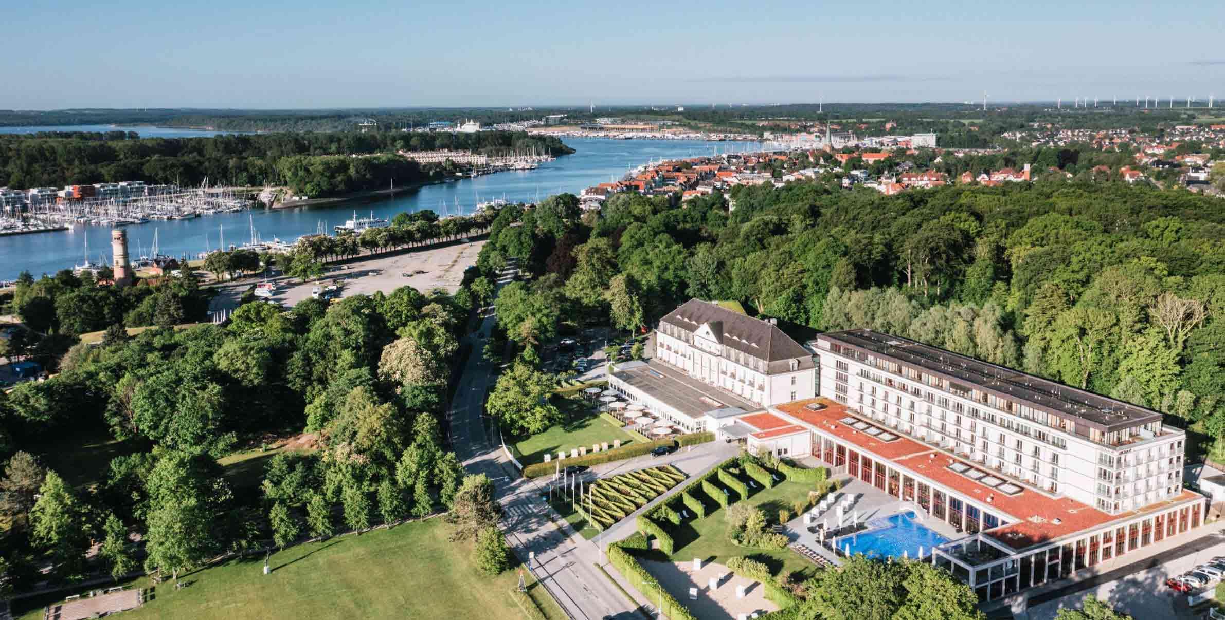 Luftbild einer Uferzone mit einer Marina voller Boote, neben einem großen Gebäude mit grünen Umgebung.