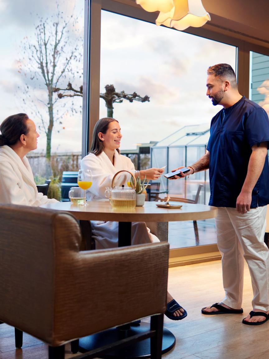 Deux clients en robes assis sur une terrasse d'hôtel, effectuant un paiement avec un lecteur de cartes portable.