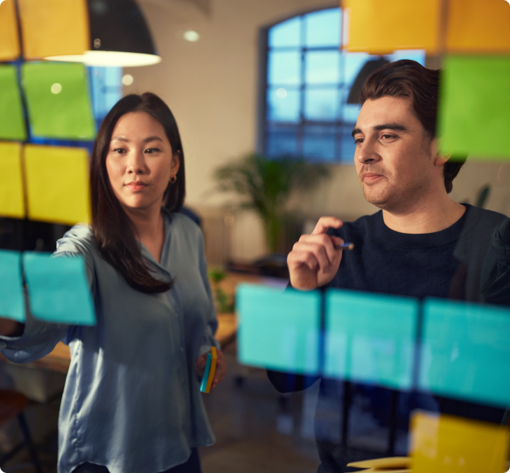 Two professionals collaborating and writing on sticky notes on a glass wall in an office setting.