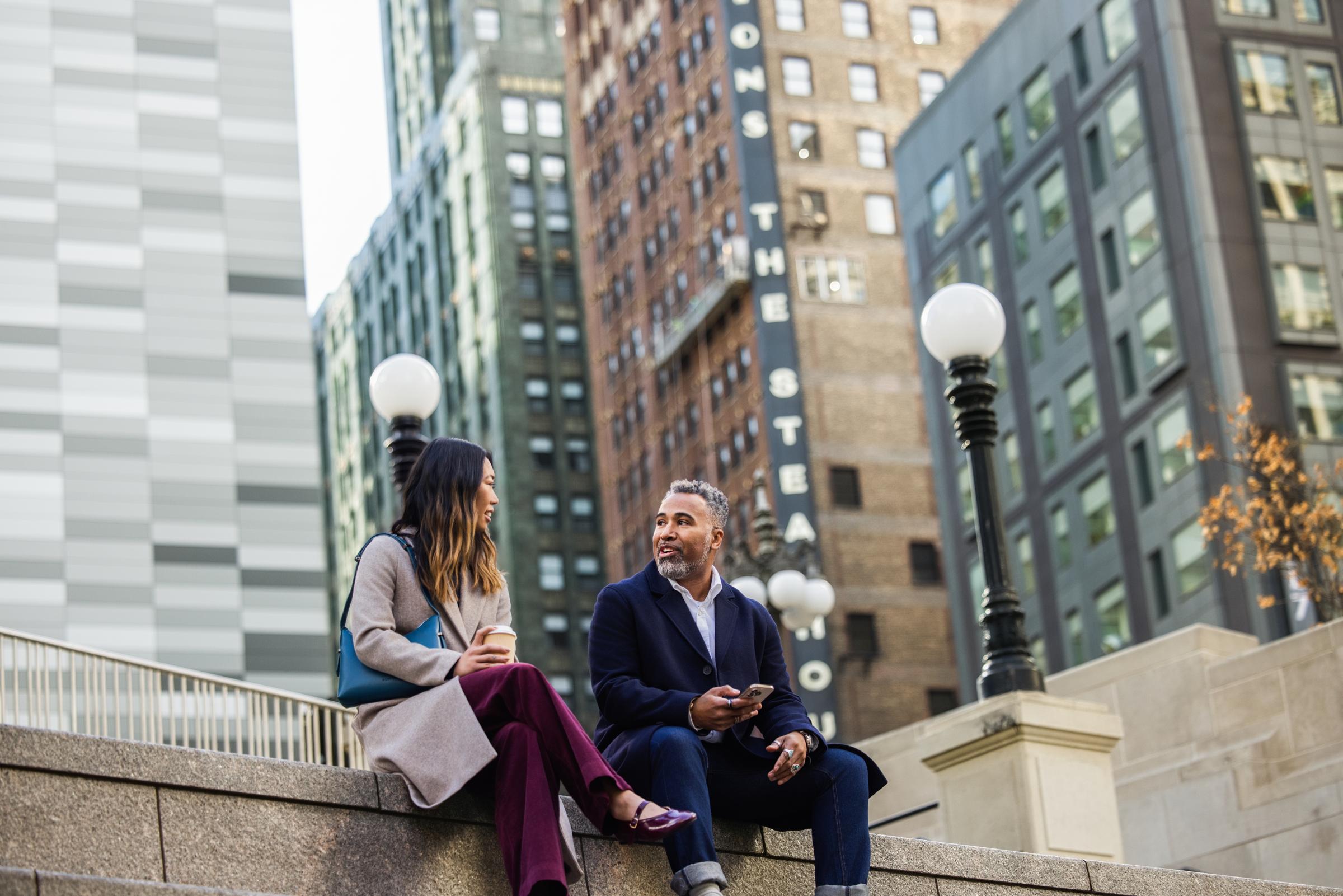 Two people sit on a city steps engaging with a mobile device near street lamps and tall buildings