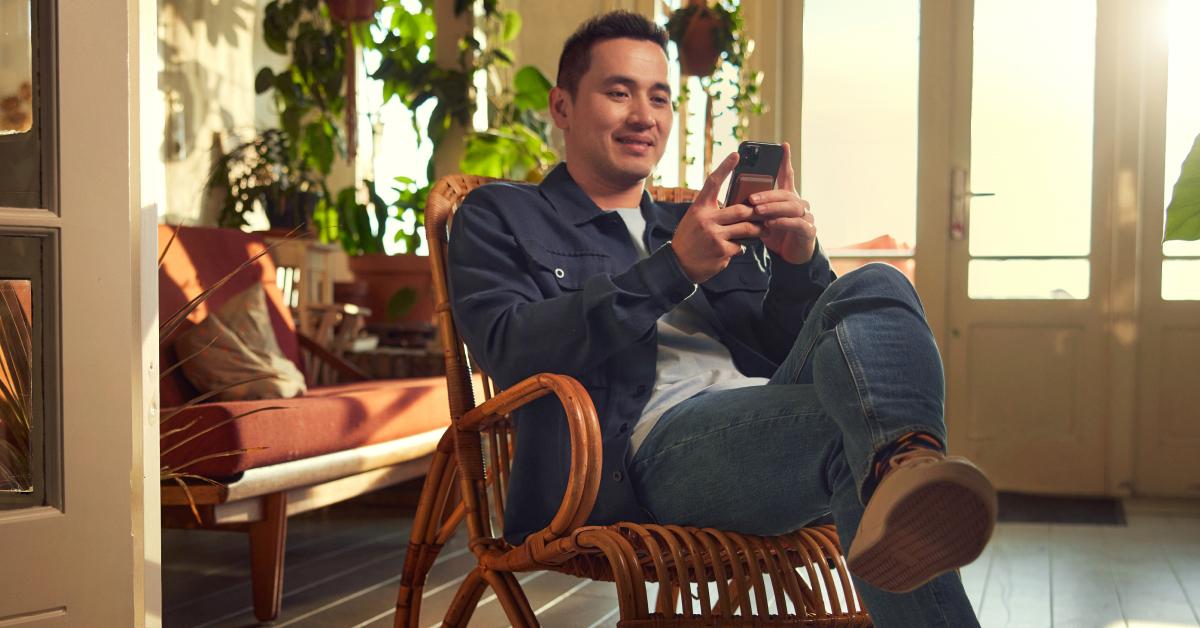Man sitting on a rattan chair using a smartphone in a cozy sunlit room.