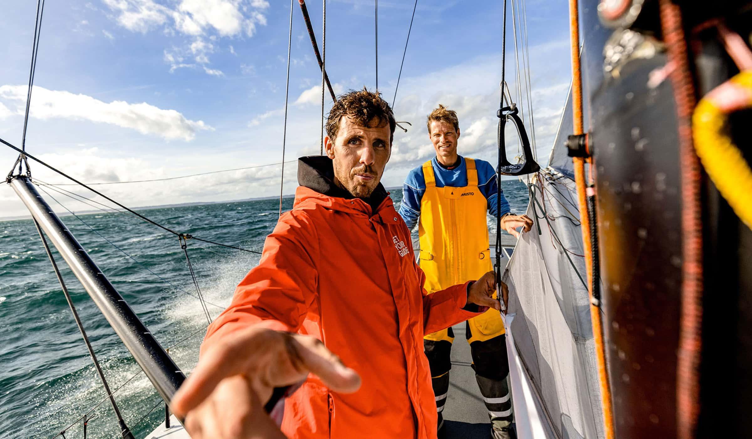Two men on a sailing boat with one extending his hand towards the camera.