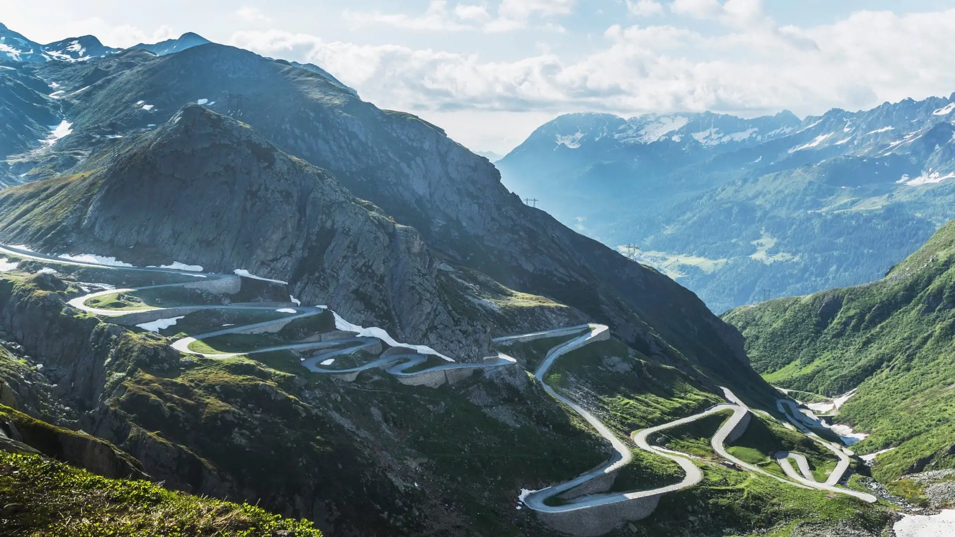 Winding mountain road with hairpin turns surrounded by green slopes and distant peaks in Switzerland.