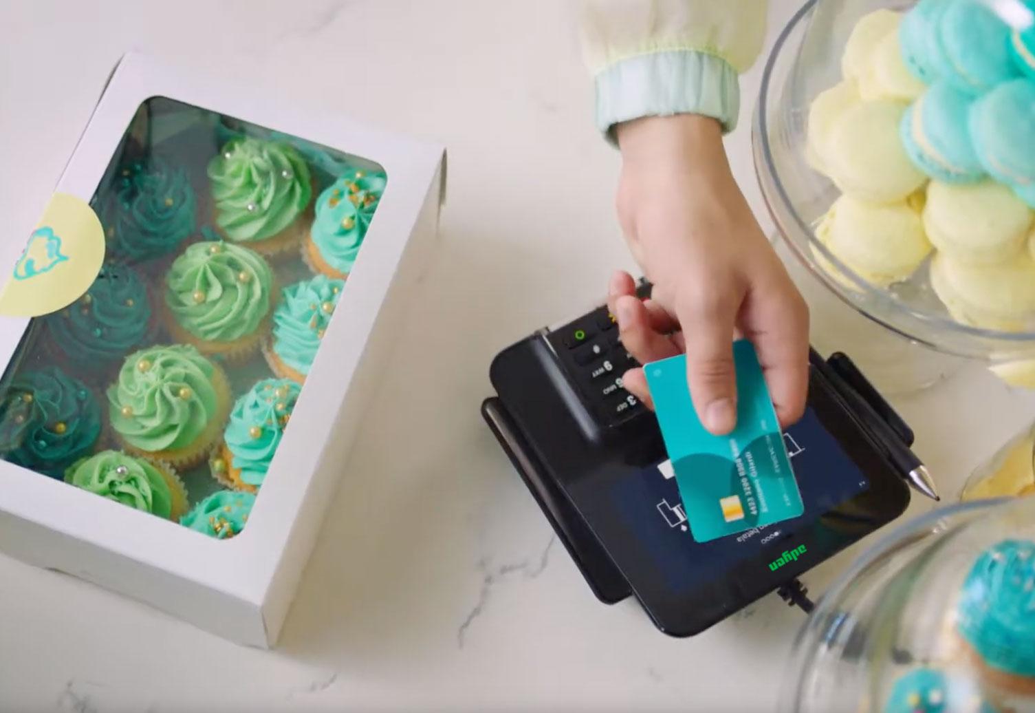 Hand, die eine Bankkarte auf einem schwarzen Adyen-Zahlungsterminal neben einer Schachtel Cupcakes streicht.
