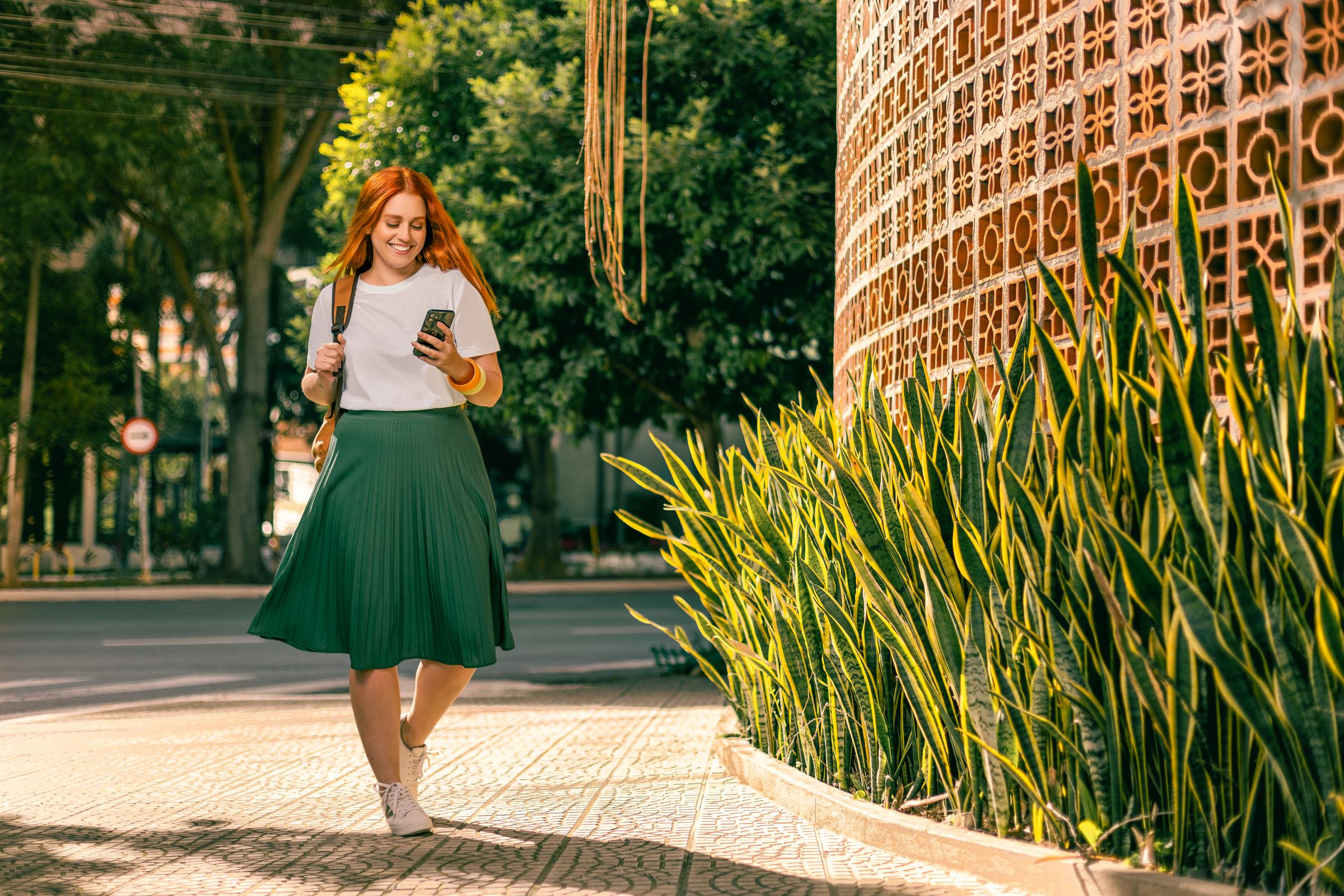 Uma mulher sorridente com uma saia verde vibrante caminhando na rua segurando um celular.