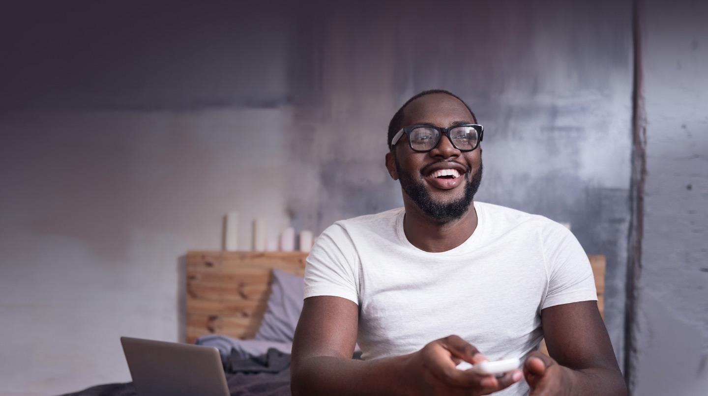 Homme souriant avec des lunettes assis sur le lit en train d'utiliser un smartphone avec un ordinateur portable à côté.