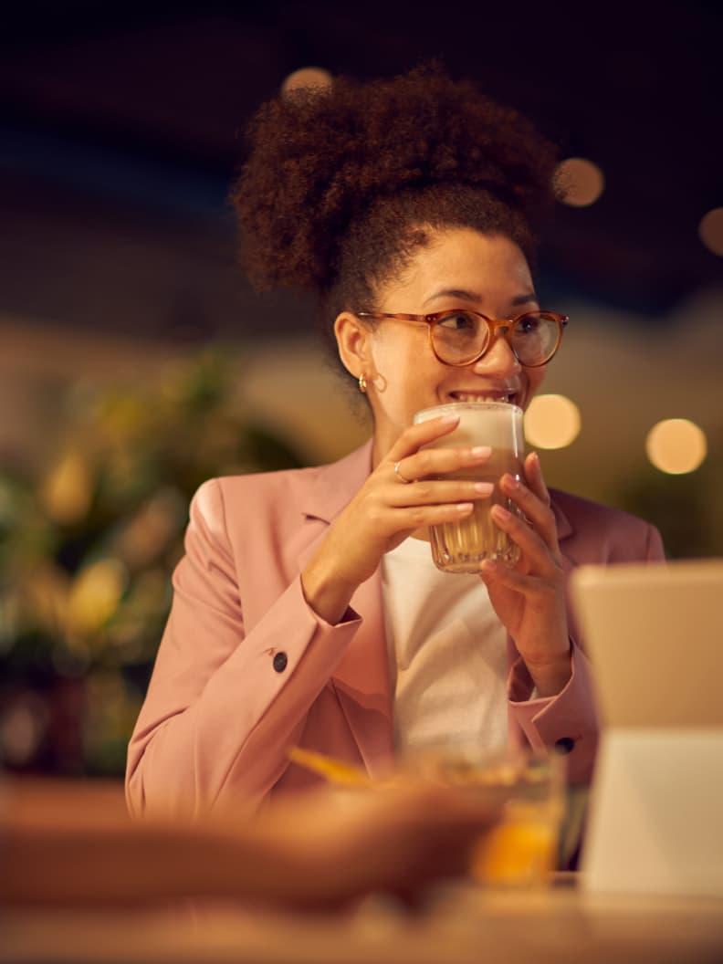 Vrouw in roze blazer drinkt een glas melk en kijkt naar laptop in een café