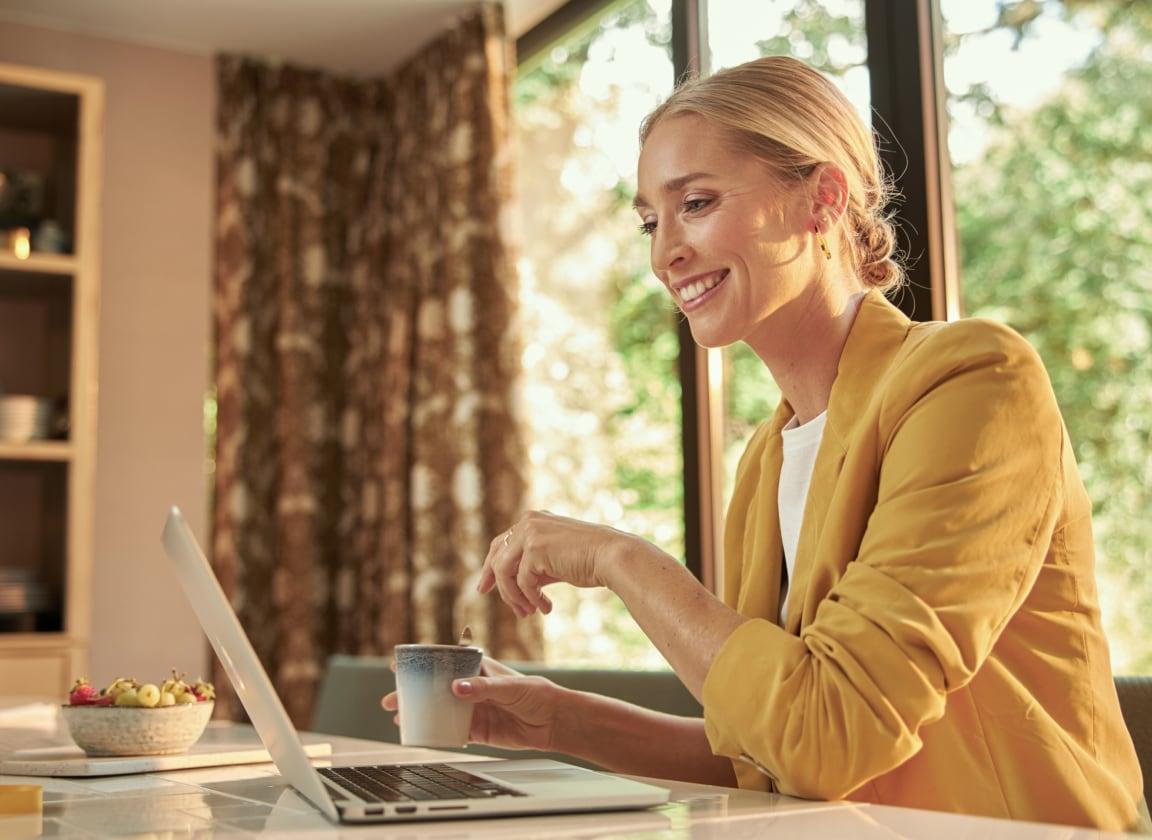 Smiling woman in a yellow jacket sitting at a table with a laptop and holding a cup.