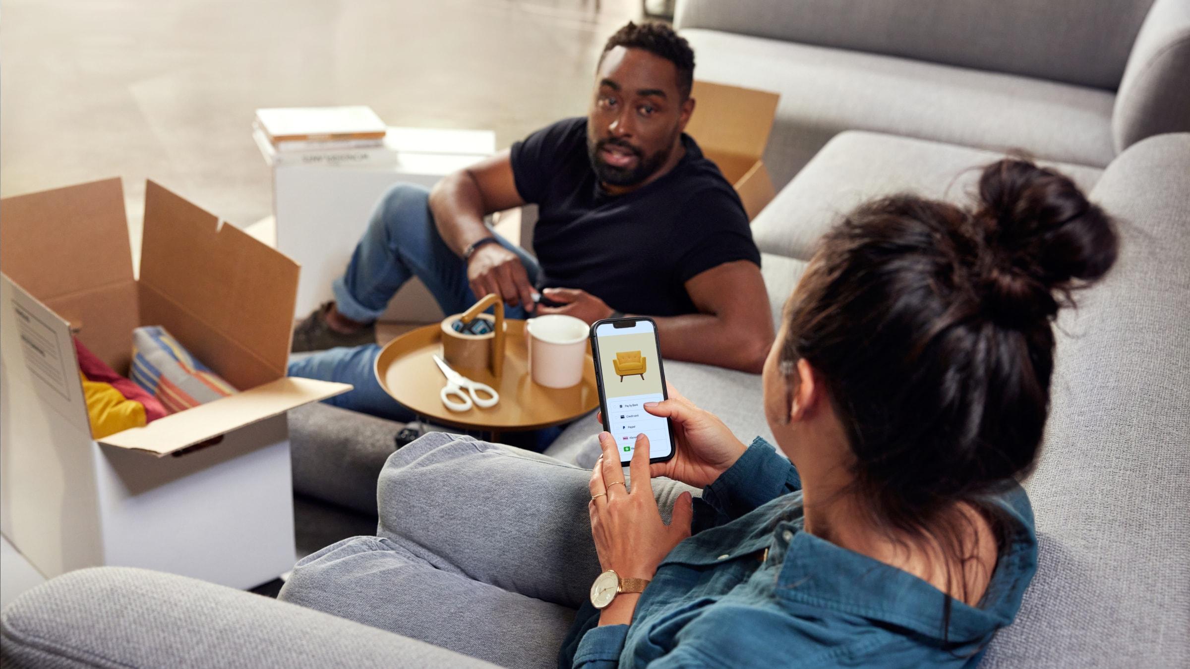 Um casal sentado no sofá e a comprar móveis no telemóvel.