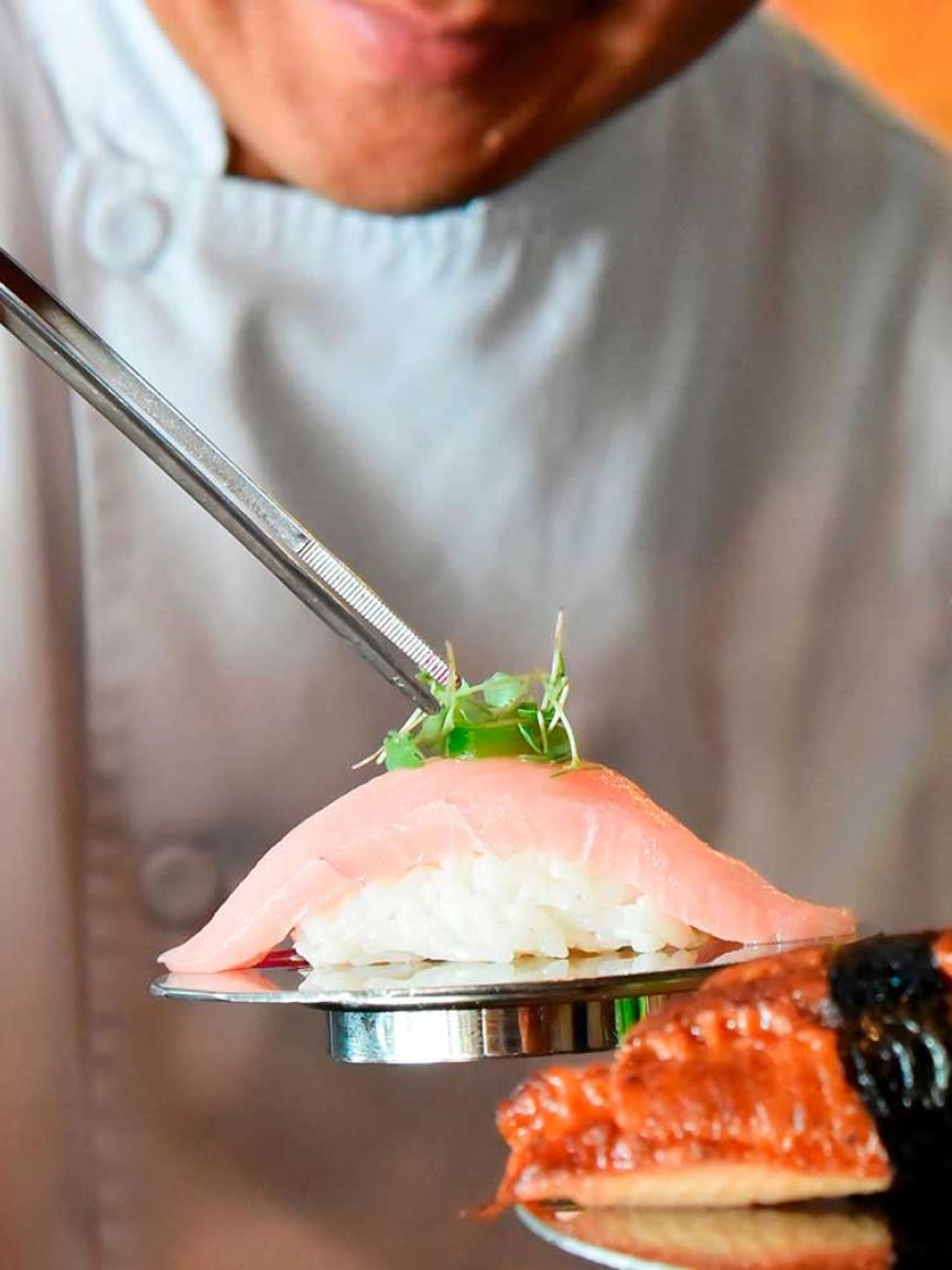 Primer plano de sushi cogido con palillos con persona desenfocada en el fondo.