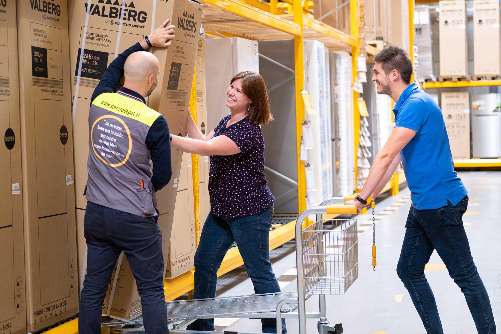 Three workers interacting in a warehouse with one pushing a trolley and another two arranging boxes.