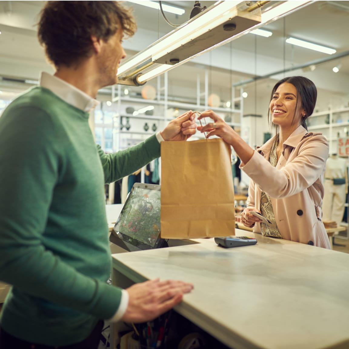 Funcionário de varejo entrega sacola de papel a cliente sorridente no caixa.