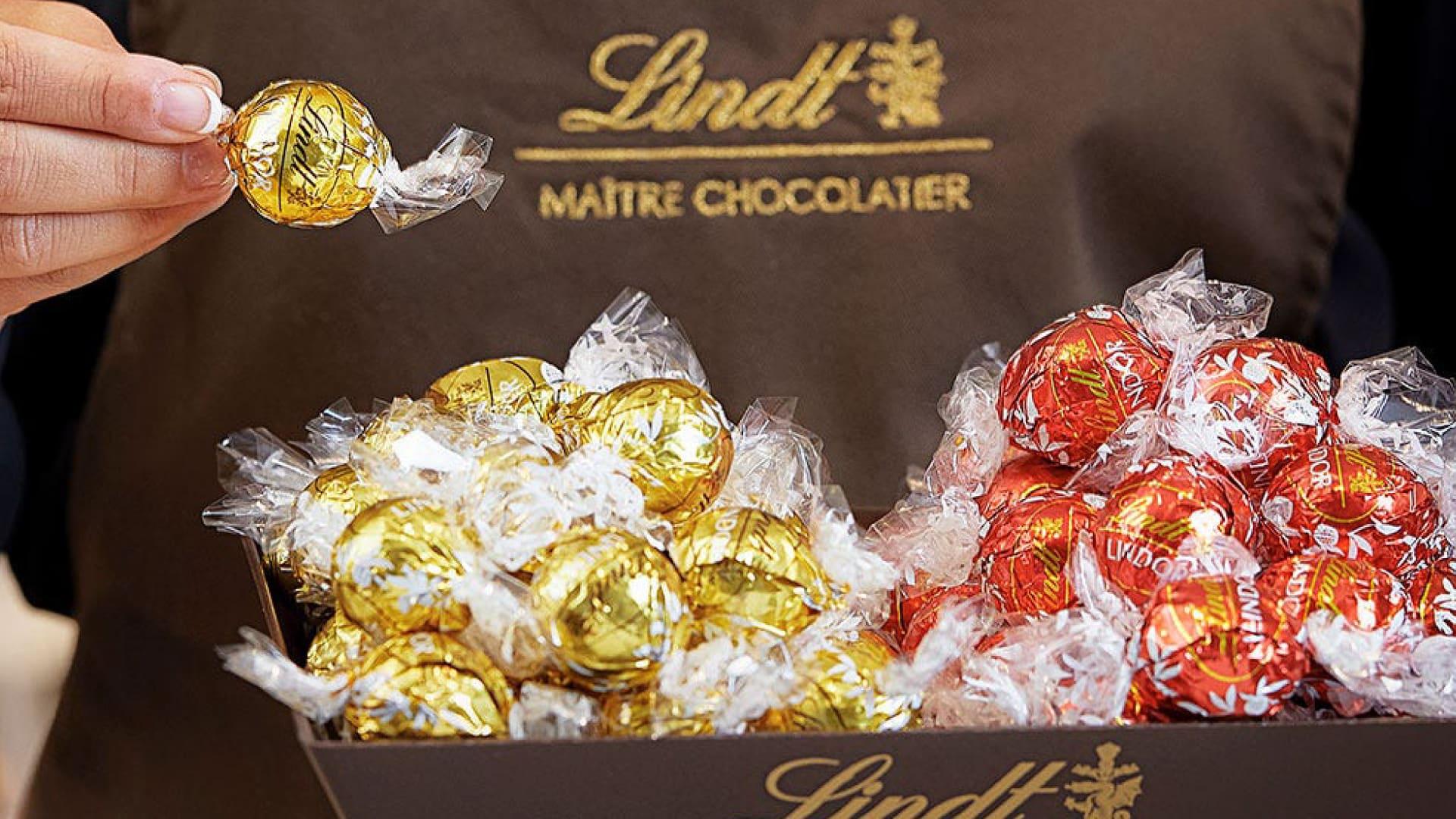 Uma pessoa segurando uma bola de chocolate Lindt embrulhada sobre uma caixa cheia de chocolates.