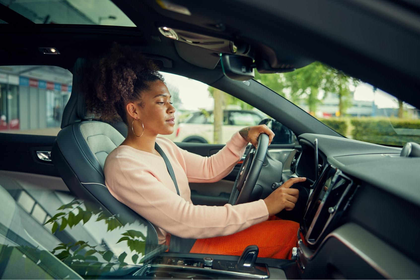 Kobieta w różowym swetrze prowadzi samochód i wchodzi w interakcje z ekranem dotykowym.