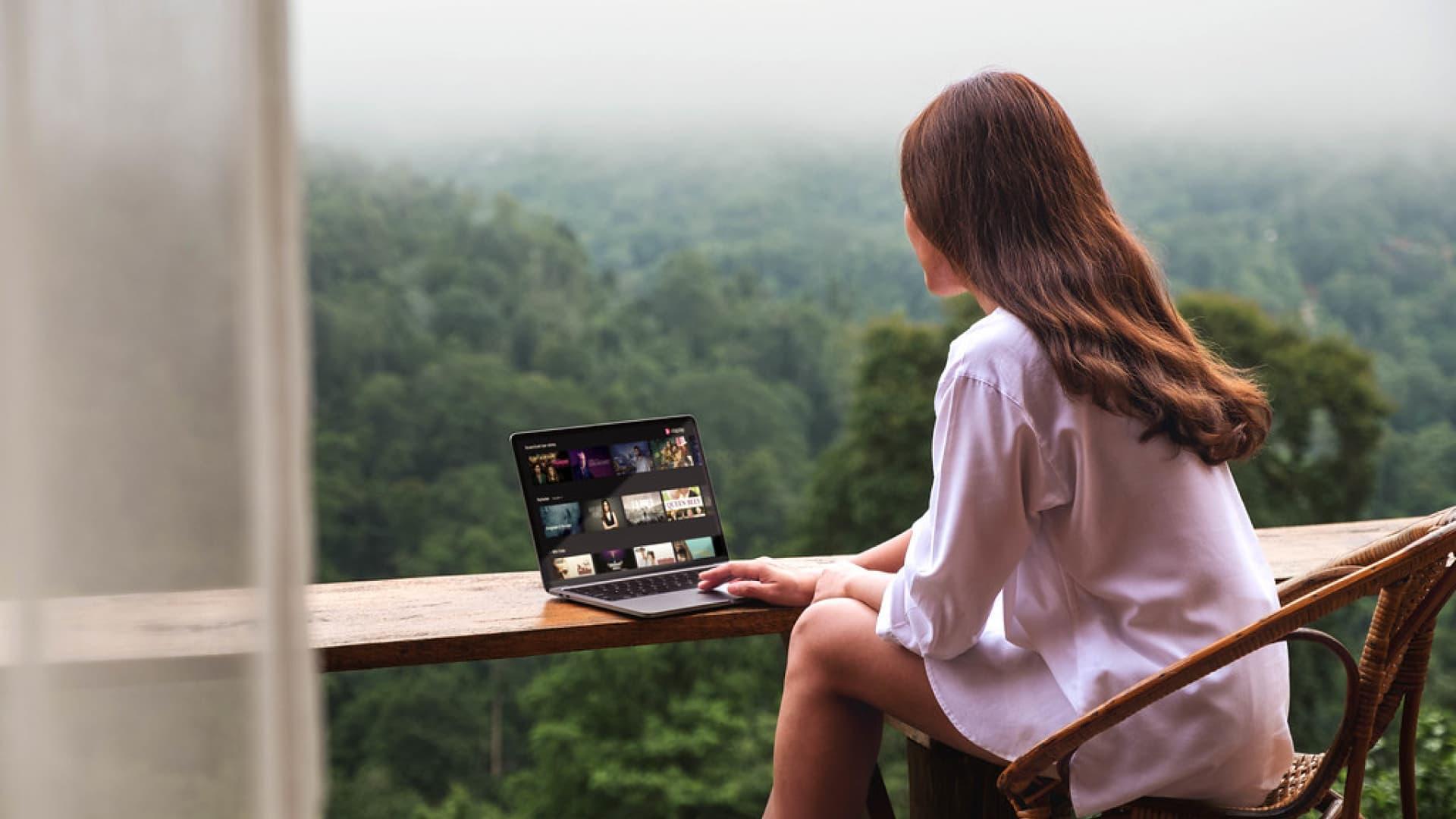 Person sittende utendørs med en bærbar datamaskin på en balkong med utsikt over en skog
