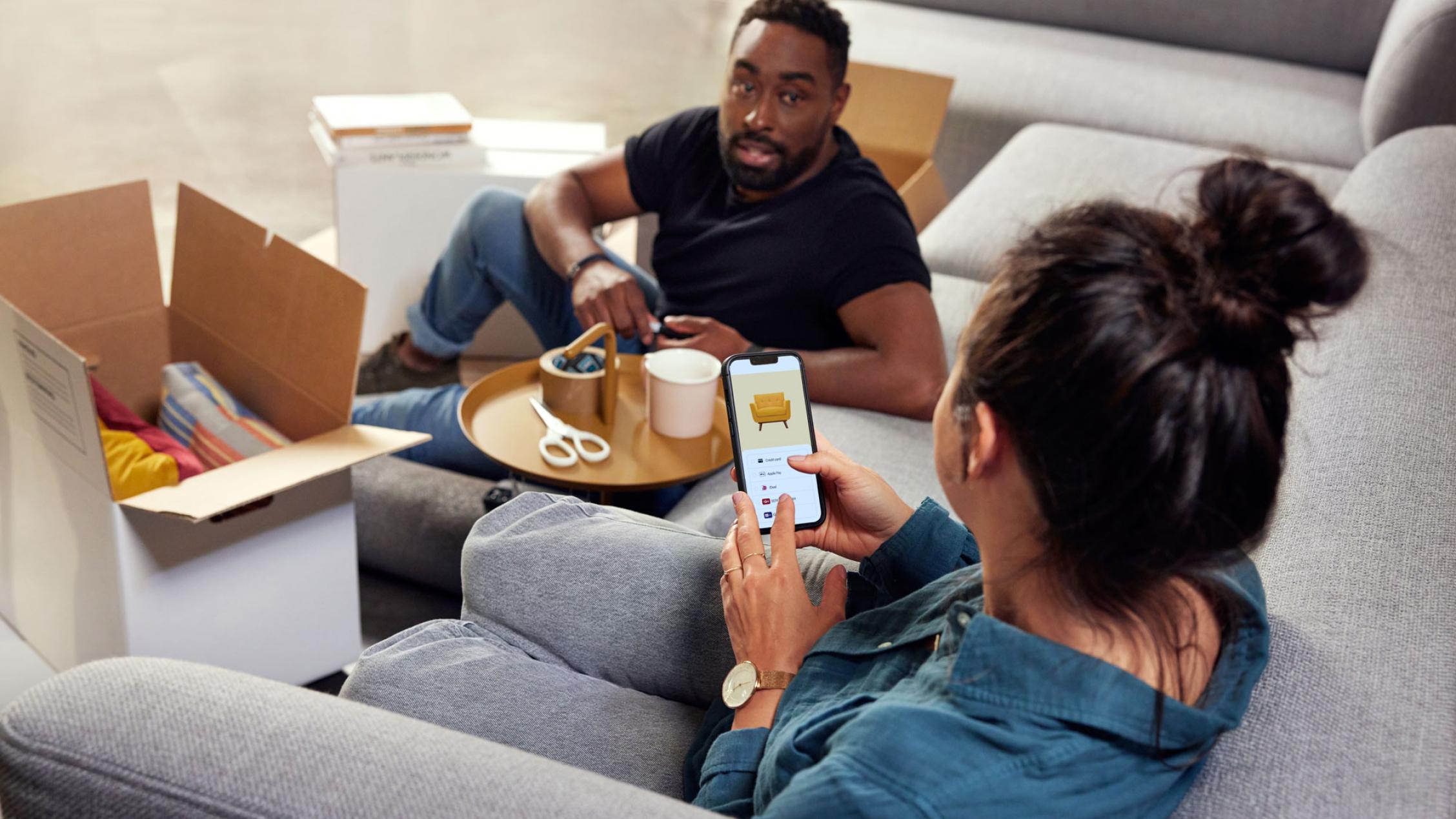 Deux personnes assises sur un canapé avec des cartons de déménagement, l'une tenant un smartphone affichant une publicité de meubles.