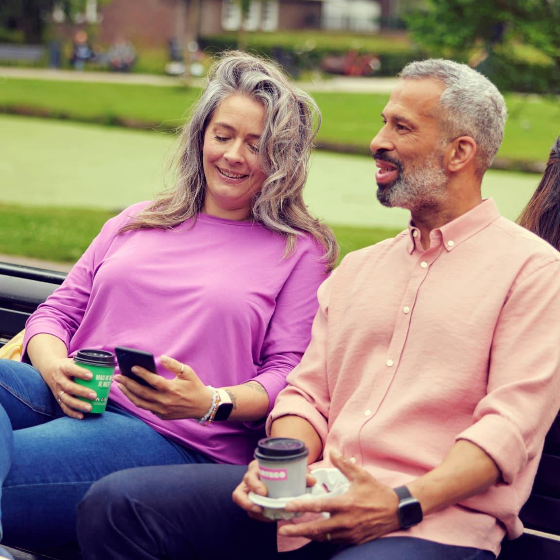 Two people sitting on a bench, holding coffee cups, and looking at a smartphone.