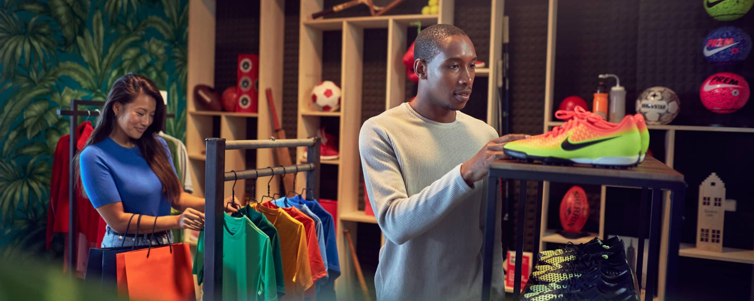 Hombre examinando zapatos deportivos en tienda con mujer sosteniendo bolsas de compras en el fondo.