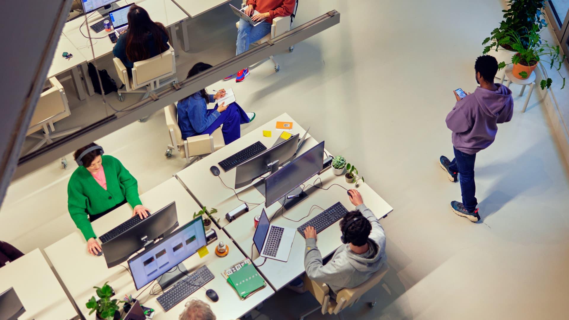 A modern office with people at desks using computers and a person walking while looking at a phone.
