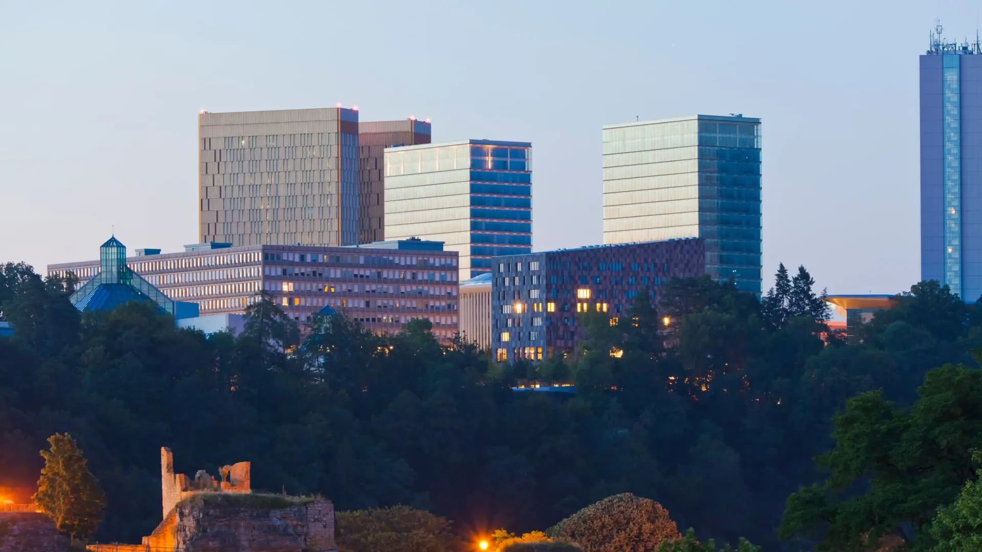 Luxembourg city skyline at twilight with illuminated buildings and greenery in the foreground.