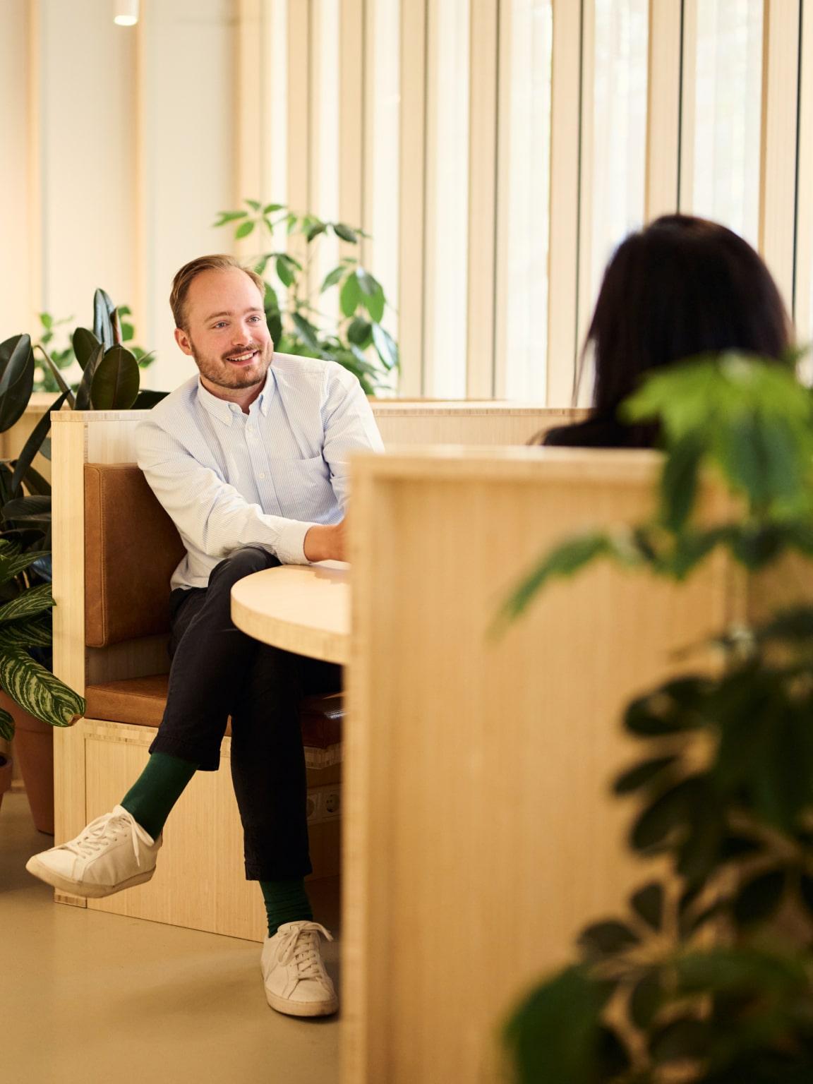 Smiling person sitting and conversing with a colleague in a bright office environment with plants.