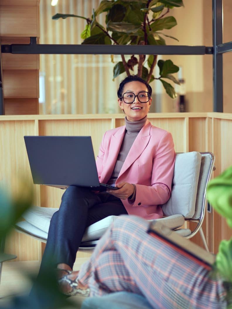Femme professionnelle portant des lunettes et une veste rose travaillant sur un ordinateur portable dans un espace de bureau moderne.