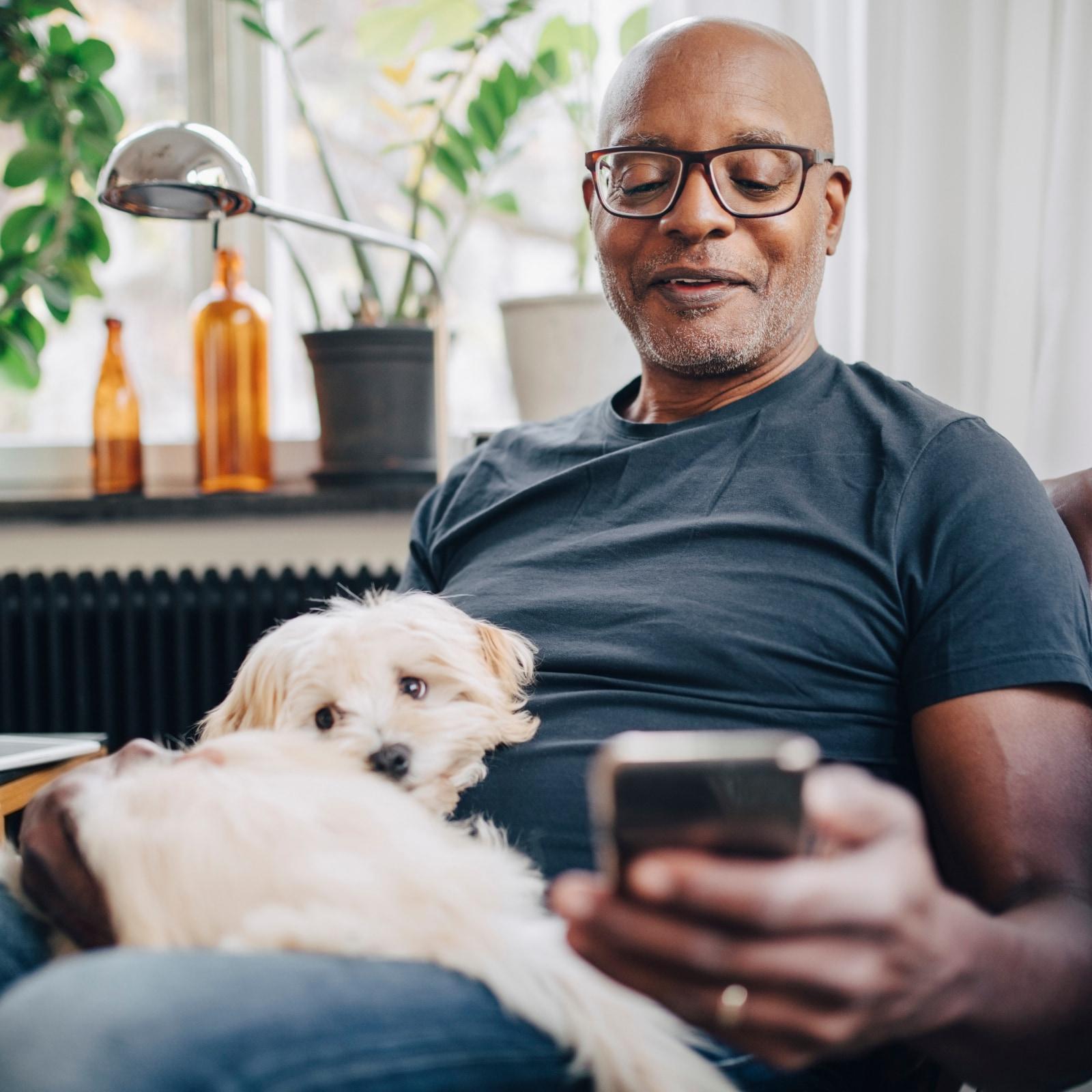 Man in glasses and a blue shirt relaxing on a couch with a small fluffy dog, looking at his smartphone.