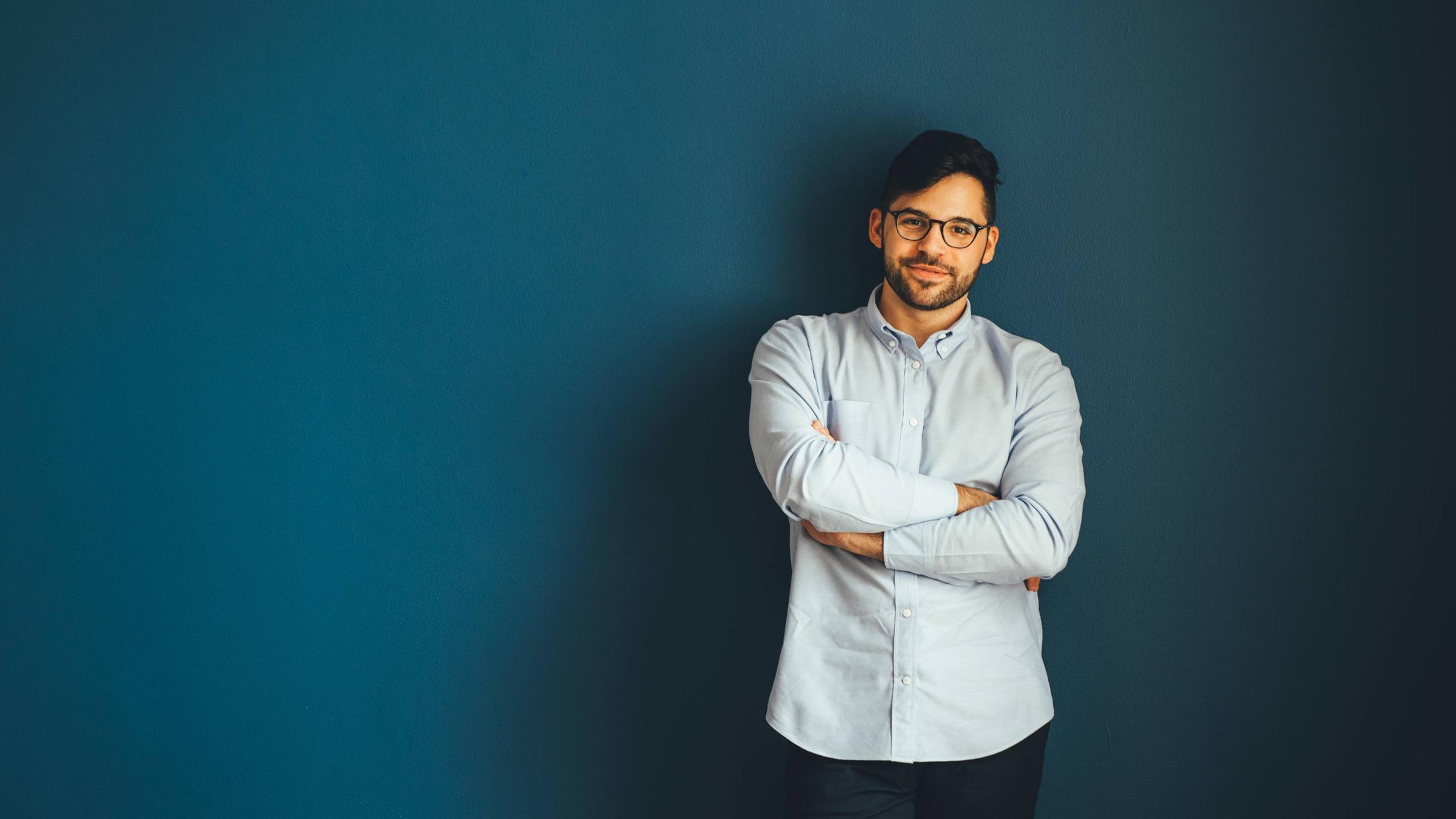 Confident man in glasses standing with arms crossed against a blue background.