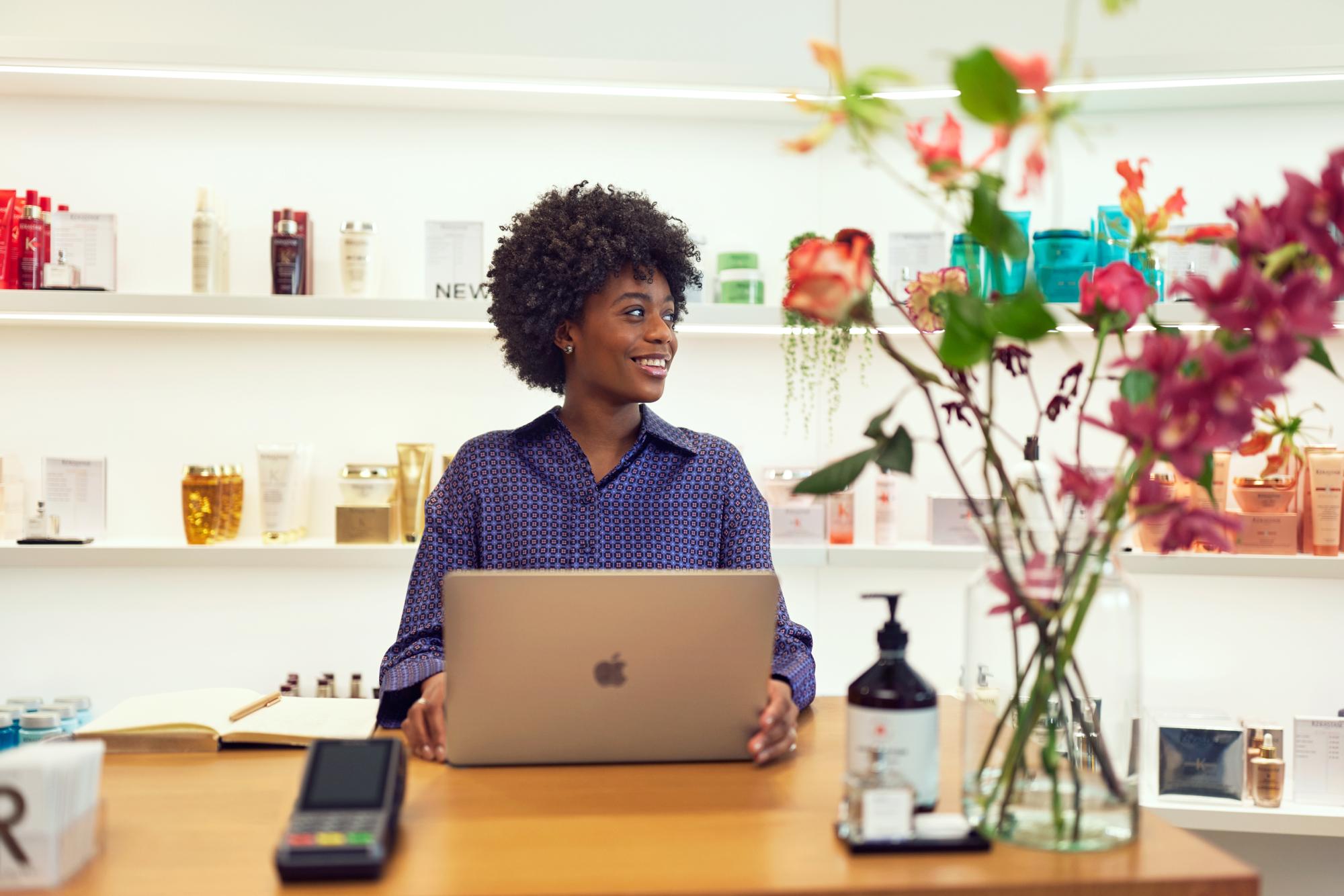 Lächelnde Person mit lockigem Haar hinter dem Tresen in einem Parfümeriegeschäft, Laptop neben einem Zahlungsterminal verwendend.