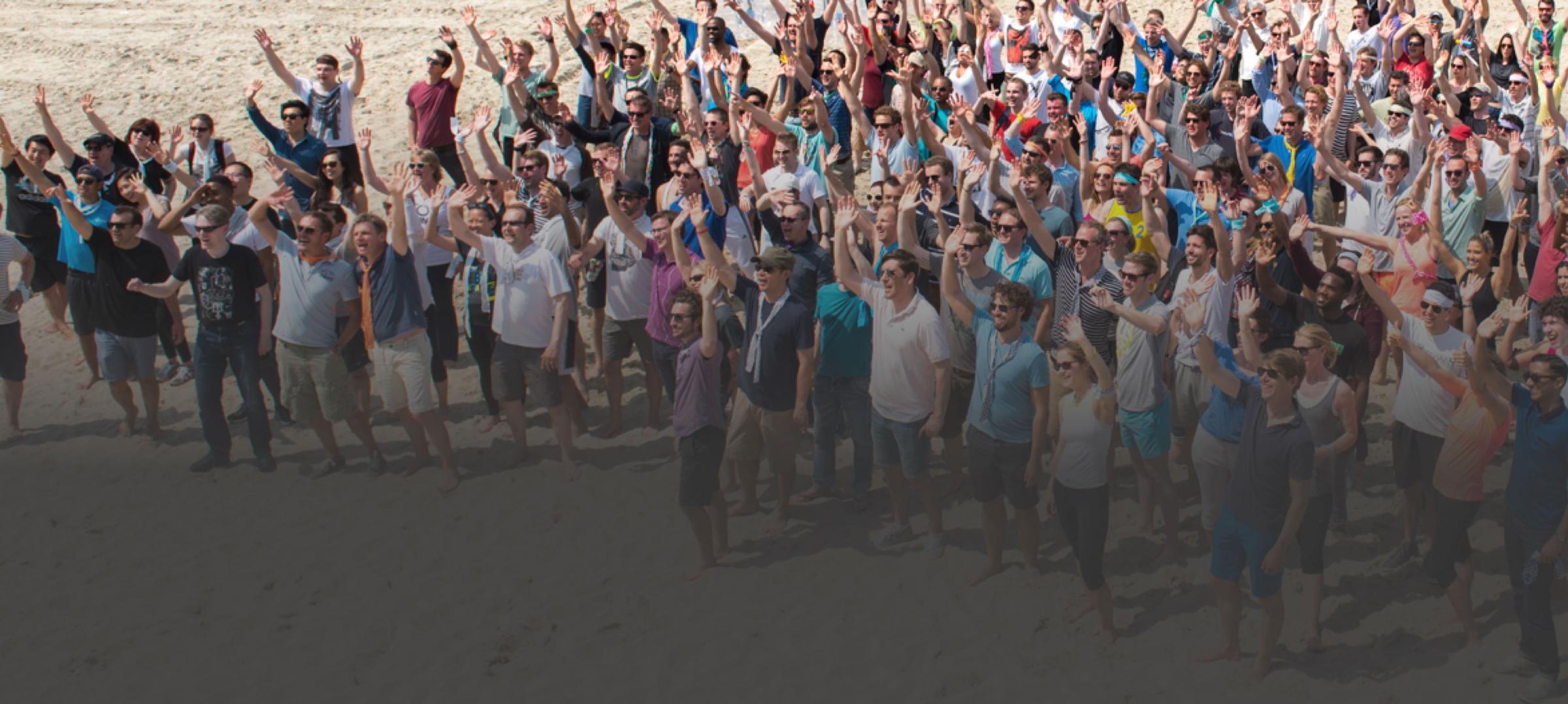 A large group of people waving and looking upwards at the camera on a sunny beach.