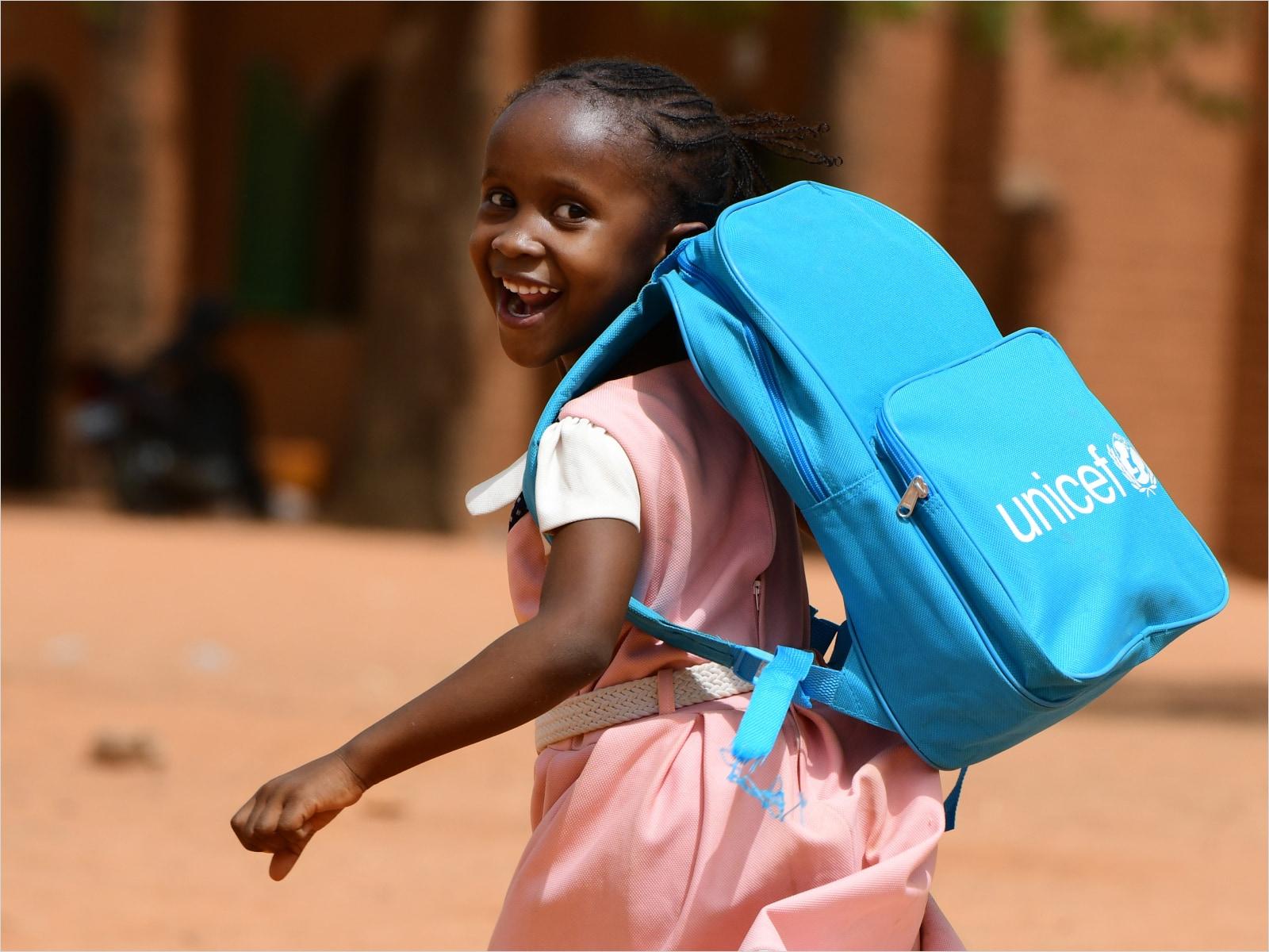 Smiling young girl with a blue UNICEF backpack walking outside.