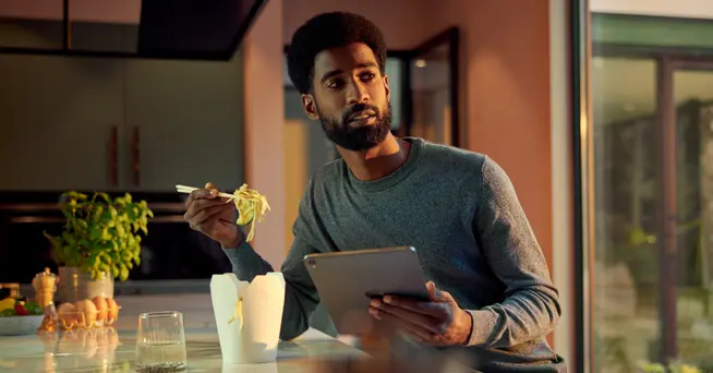 Homem comendo noodles e olhando para um tablet em uma cozinha moderna ao entardecer.