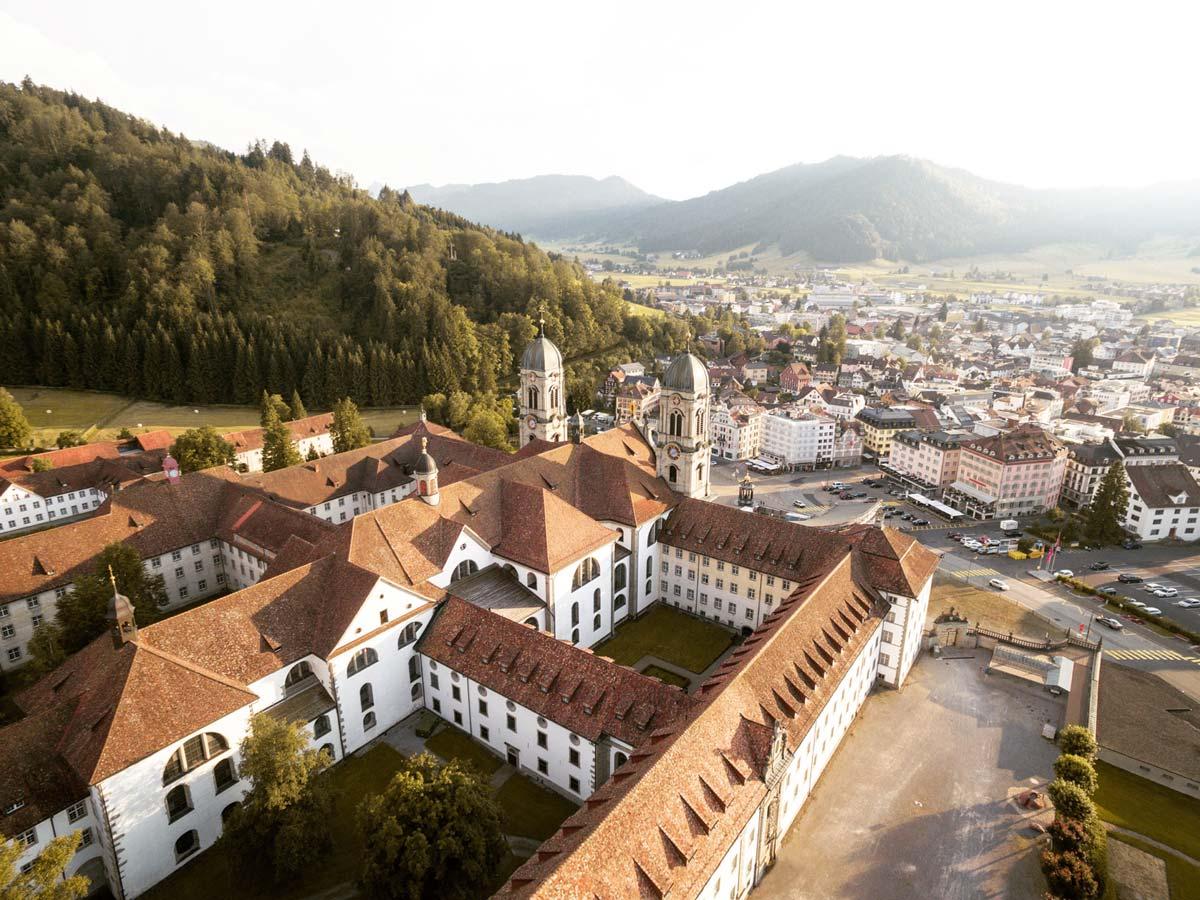Luftbild einer historischen Stadt mit Klosterkomplex und umliegenden grünen Hügeln.
