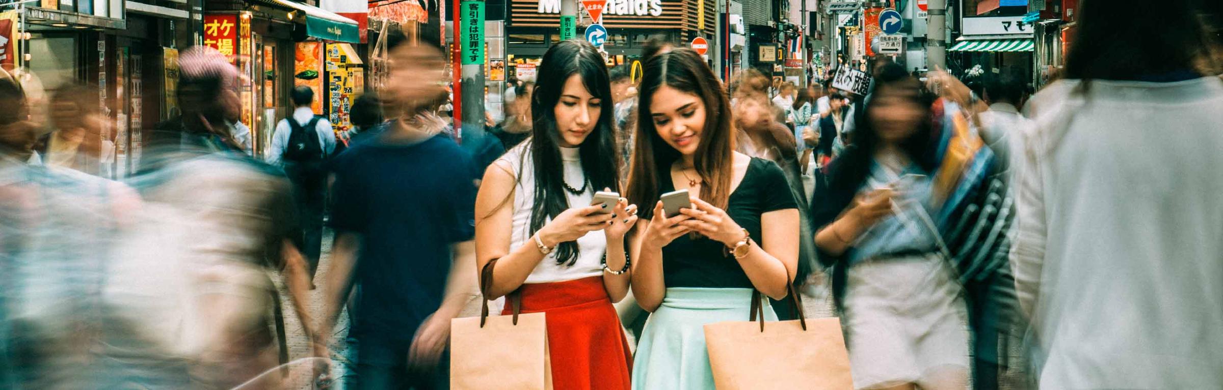 Deux femmes utilisant des smartphones tandis qu'elles se tiennent debout dans une rue animée avec des passants flous.