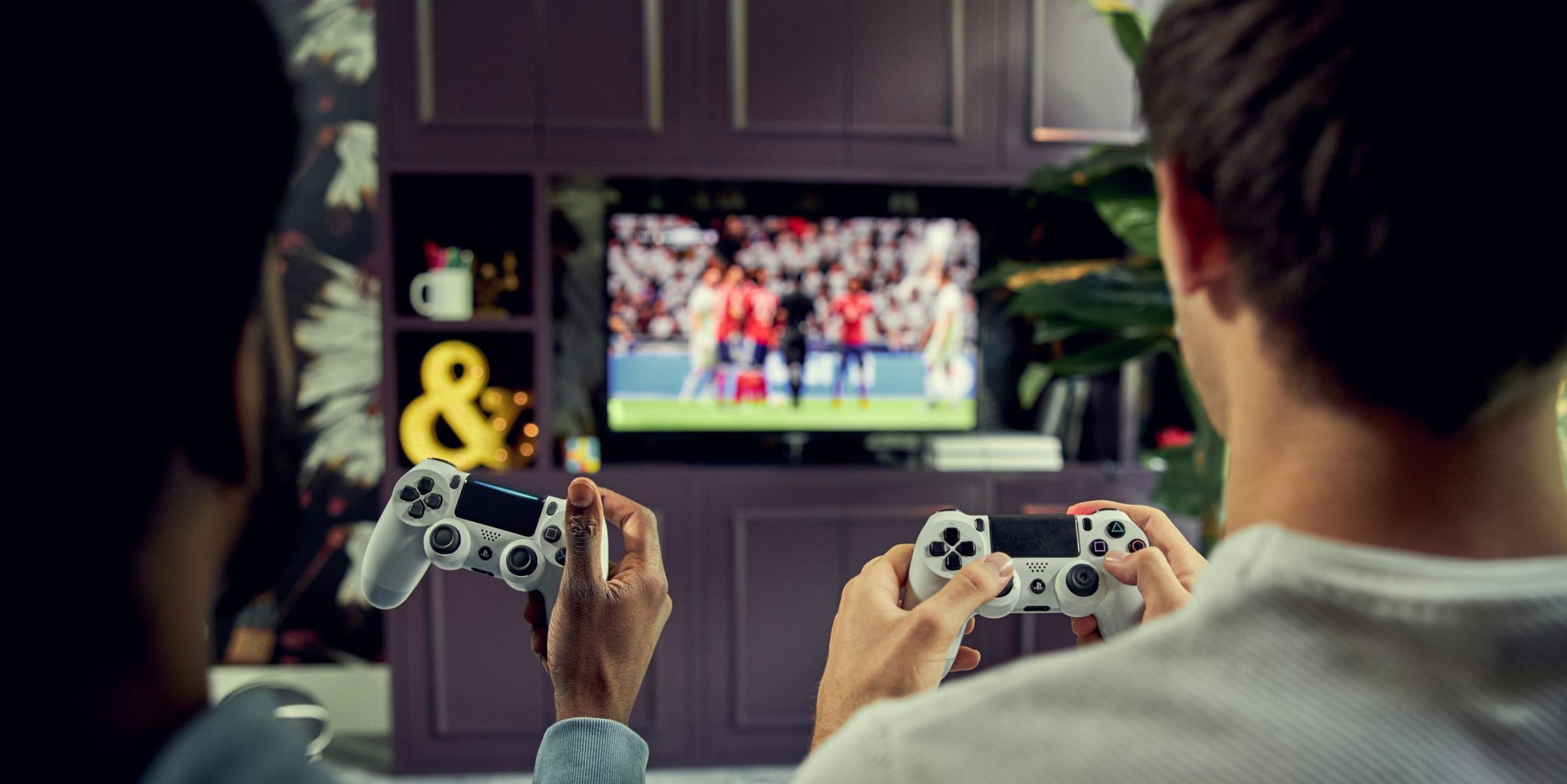 Two people playing a soccer video game on a TV screen from an over-the-shoulder perspective.