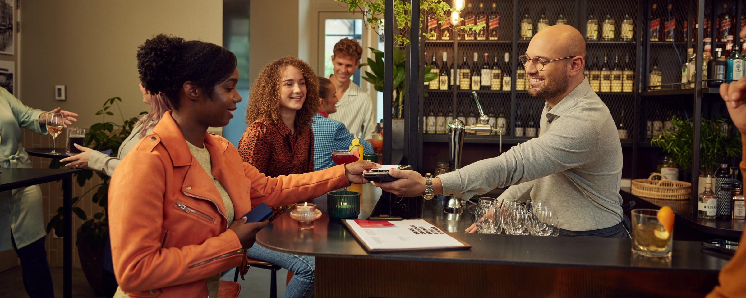 Clientes disfrutando de bebidas mientras un bartender procesa un pago con un terminal de Adyen.