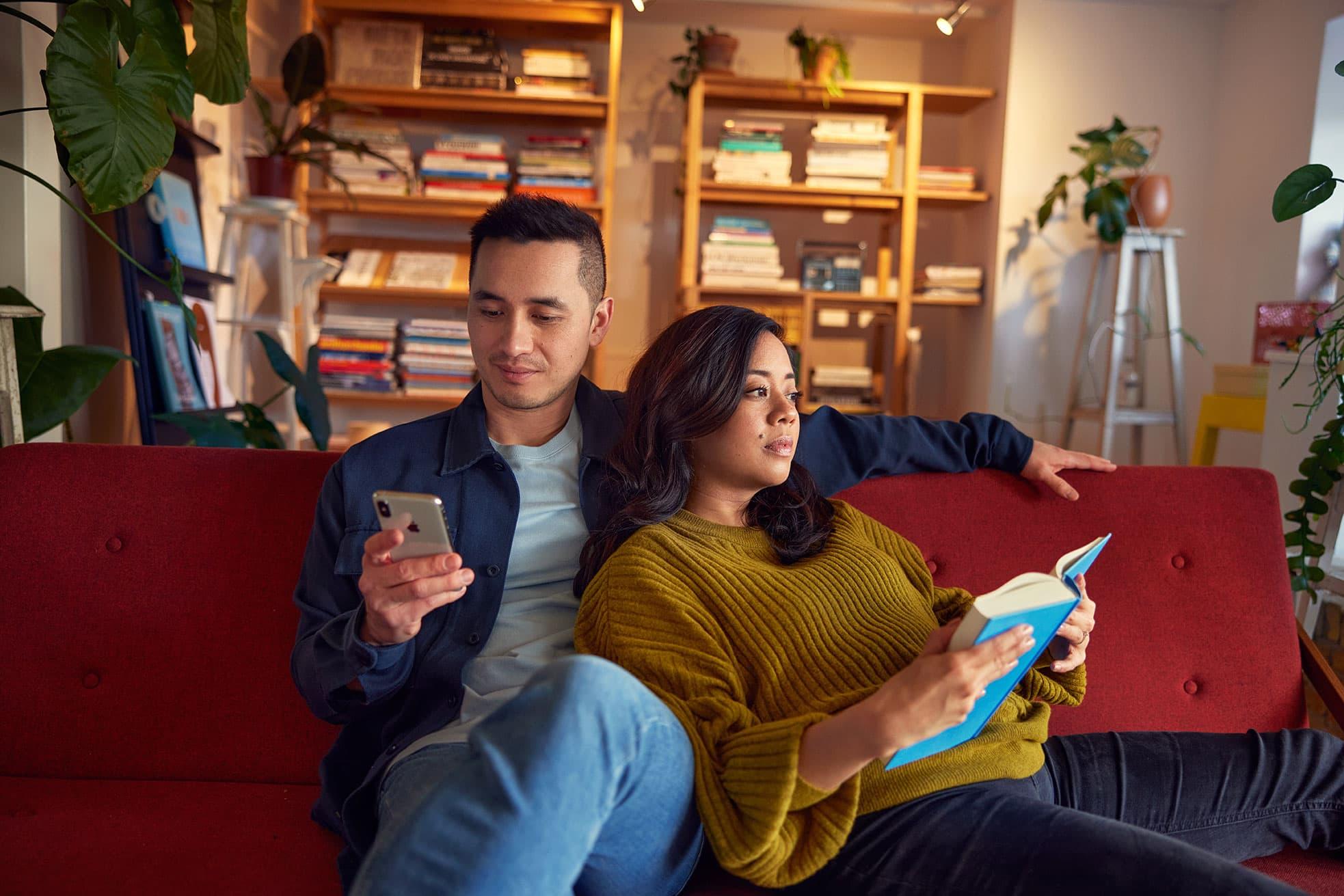 Homem usando smartphone e mulher lendo um livro em um sofá vermelho em uma sala de estar aconchegante.