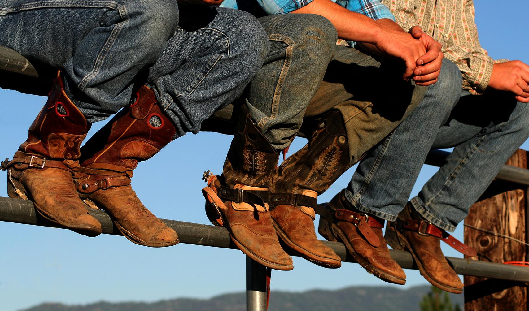 Three individuals wearing cowboy boots sit on a fence in a rural setting.