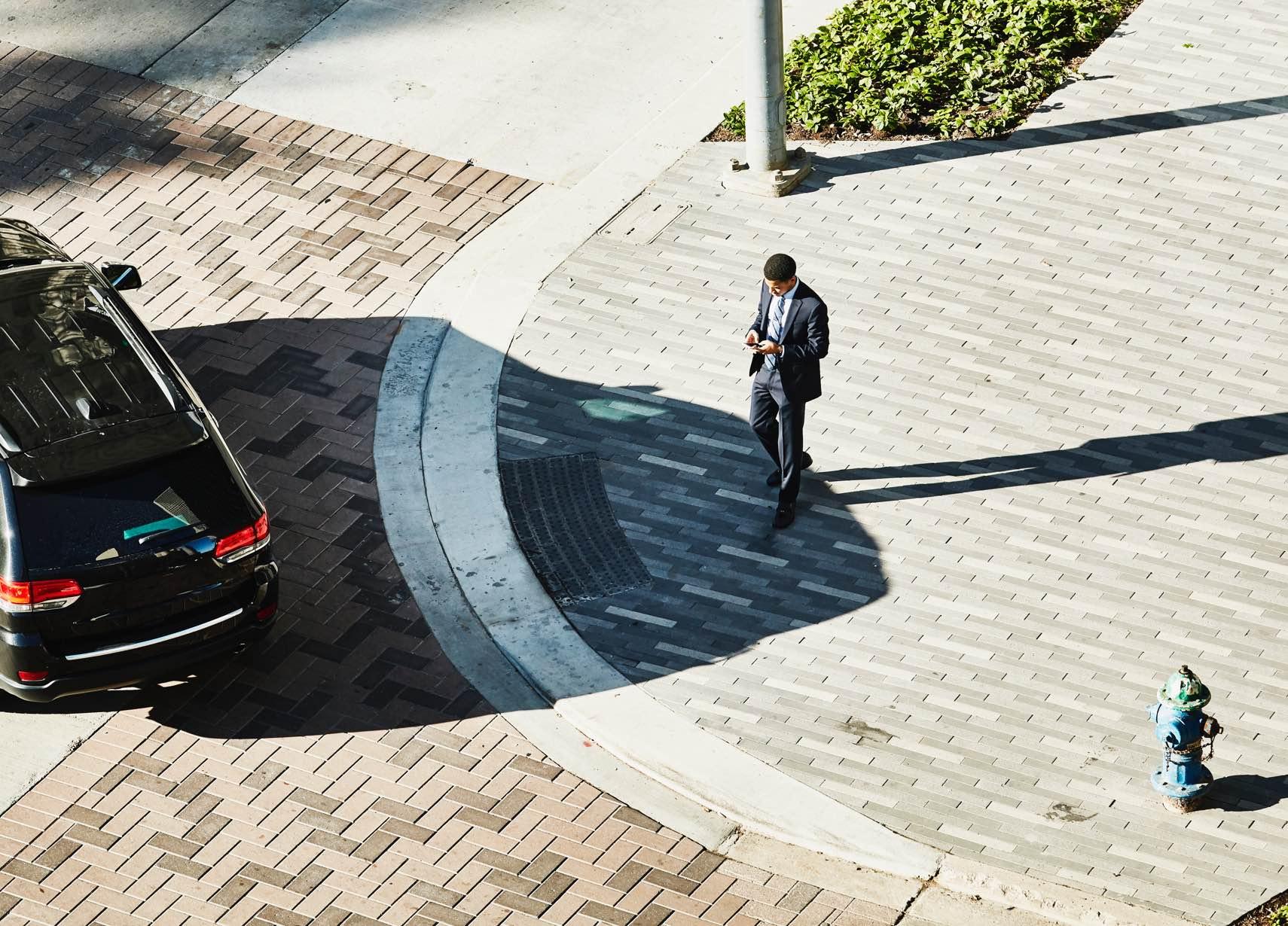 Vista aérea de um homem de terno caminhando em uma calçada com padrão, ao lado de um carro estacionado.