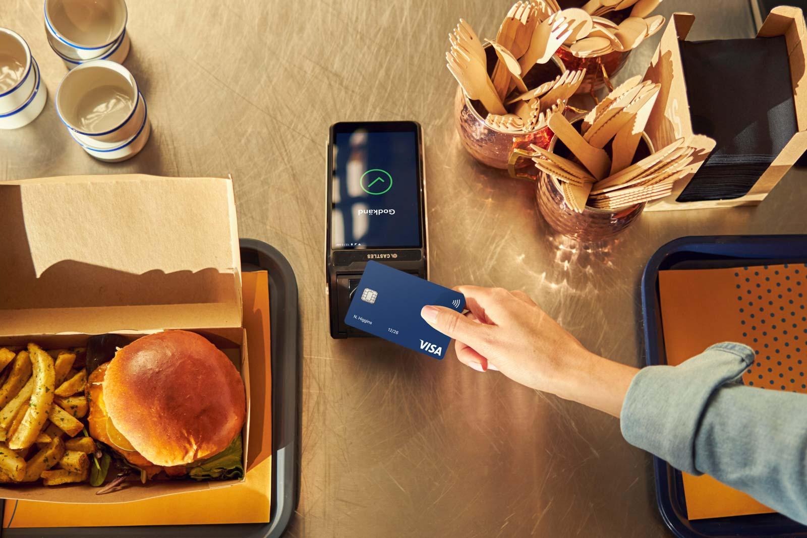 Person som använder ett kontaktlöst Visa-kort på en Adyen-betaltjänst med en hamburgarmåltid bredvid.