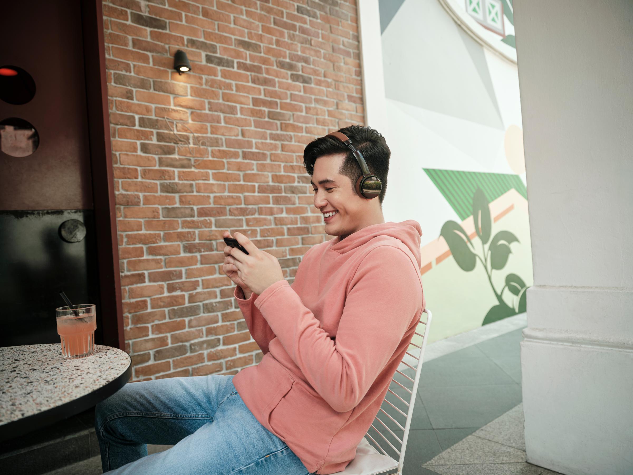 Um homem sentado à mesa, olhando para o telefone, com fones de ouvido e rindo