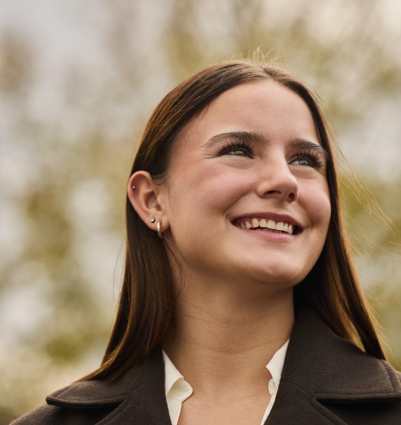 Sienna Gschwind in dunklem Mantel vor Naturhintergrund bei Tageslicht in der Untersichtsperspektive.