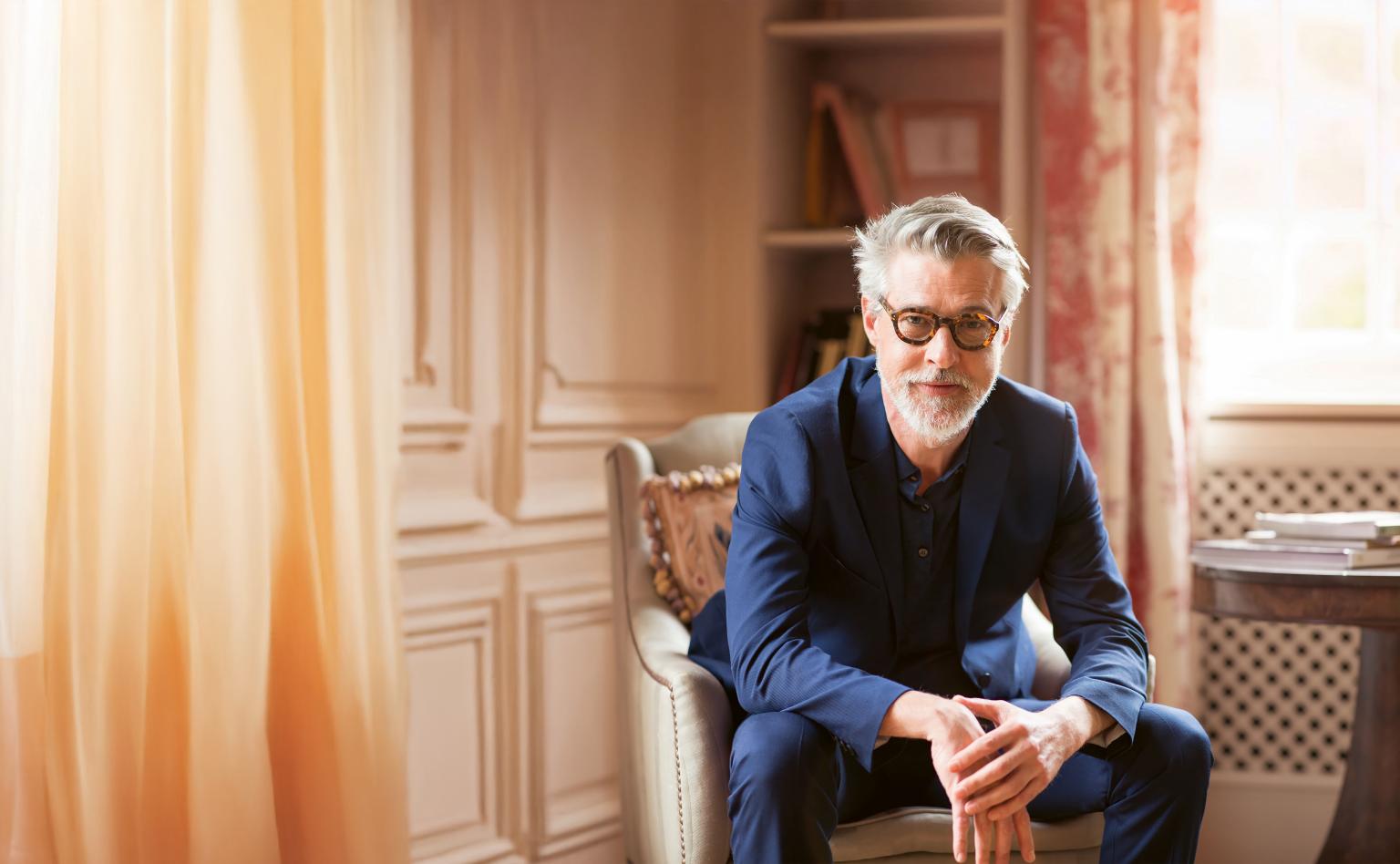 Ein Mann im Anzug sitzt in einem gemütlichen Raum neben einem Tisch mit Papieren.