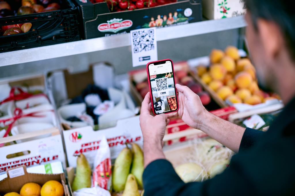 Eine Person scannt mit dem Smartphone einen QR-Code auf einem Obststand.