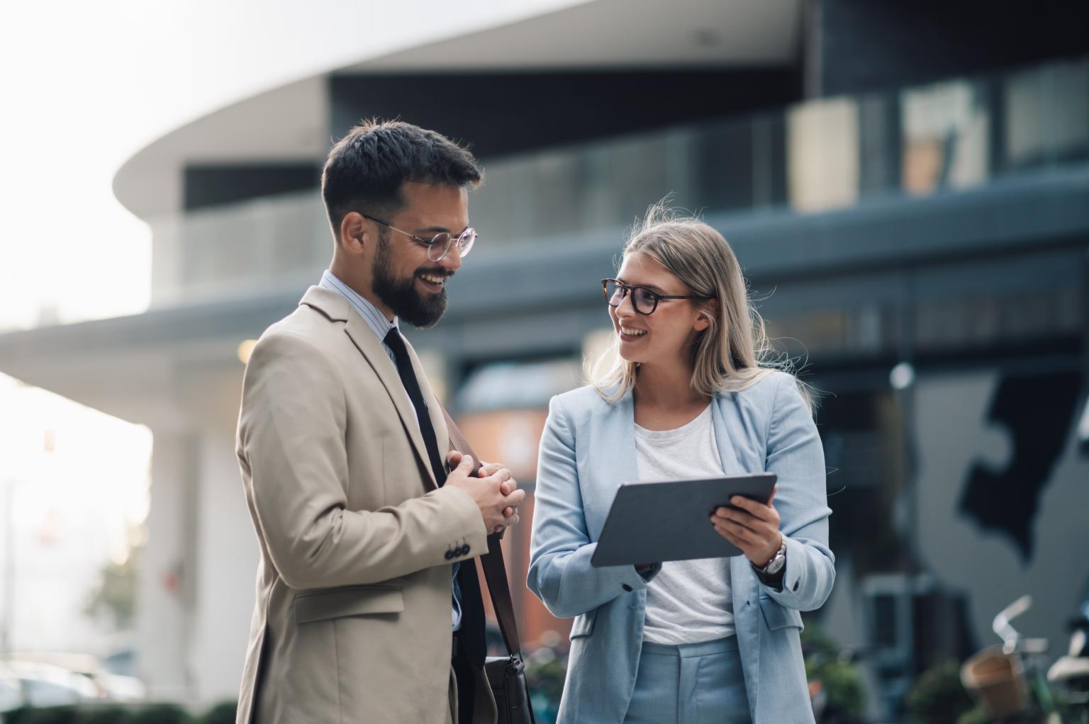 Two businesspeople are having a conversation outdoors on a sunny day, smiling and looking at each other while one holds a digital tablet